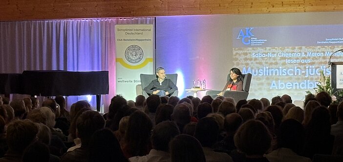 Blick in den Vortragssaal mit Publikum von hinten und Bühne mit zwei Personen in Sesseln sitzend. 